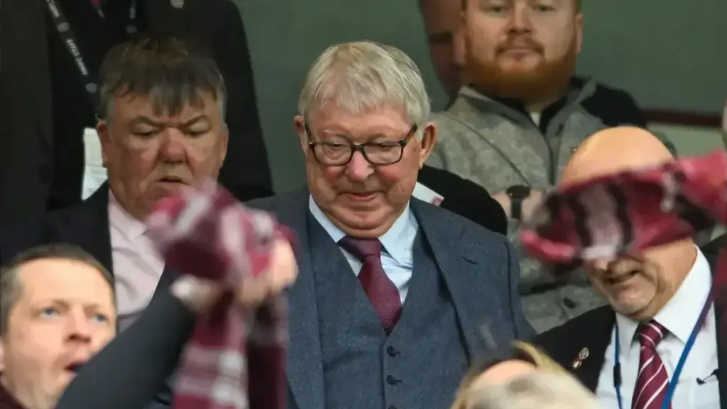 Sir Alex Ferguson seated in the stands at a football match, wearing a suit and glasses, with several spectators around him.